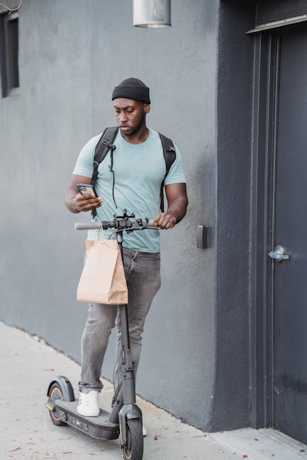A young man with dark skin, wearing a black beanie, light blue t-shirt, and gray jeans, stands outside against a gray concrete wall. He is holding a smartphone in his right hand and a paper bag in his
