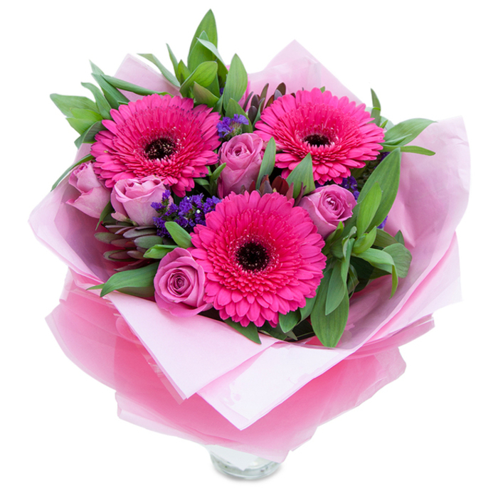 I've always loved how pinks can balance playfulness and quiet grace, and this hand-curated bouquet demonstrates that balance clearly. Three exuberant Gerbera daisies in vivid fuchsia hold the eye with their layered petals and dark centres, while a handful of lavender-tinged roses unwrap inwards like soft secrets, petals overlapping in a delicate spiral. Between them sit tiny clusters of deep purple statice, adding texture like little wildflower memories tucked into a formal arrangement. The greenery behind - glossy, broad leaves similar to salal or eucalyptus - gives the bouquet weight and a cool green contrast that makes the pinks sing. The flowers are cocooned in a pale pink tissue lining with an outer sheet of sheer fuchsia cellophane that shimmers faintly; the wrap is folded with the sort of care you'd expect from a local florist packing flowers for pick-up near Wanstead Flats or for a delivery to a flat off Upton Lane. The lower edge reveals the top rim of a clear glass vase, so the bouquet arrives ready to stand on a table and catch the light. Under soft afternoon light, the velvety roses offer a subtle perfume while the Gerberas contribute a fresher, green scent. This arrangement feels intimate and celebratory at once - ideal for marking anniversaries, birthdays, or simply sending an affectionate 'you're special' to someone across Forest Gate and neighbouring streets.